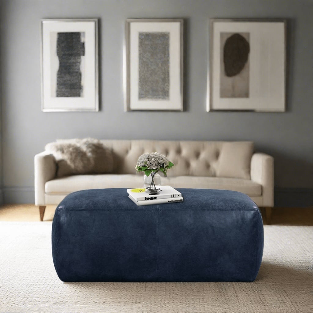 A modern living room featuring a large blue leather ottoman in the center, a beige sofa with a plush throw, and three abstract black and white framed artworks on a light grey wall.