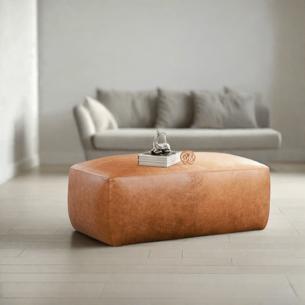 A minimalistic setting showcasing a large tan leather ottoman on a pale wooden floor, with a modern sofa in the background, and a small sculpture on top of a book resting on the ottoman