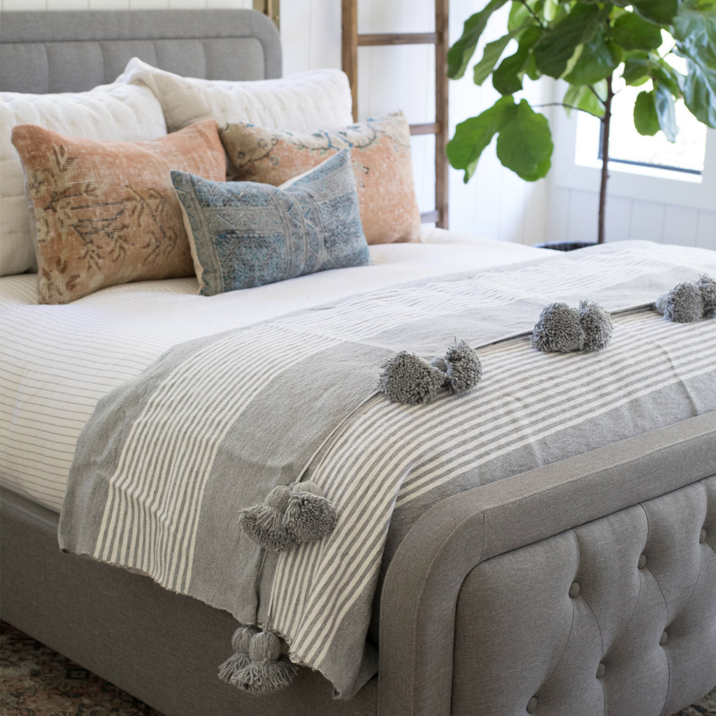 Gray Stripes Moroccan Pom pom blanket on top of a bed with pillows in a bedroom with furniture - Moroccan Interior 