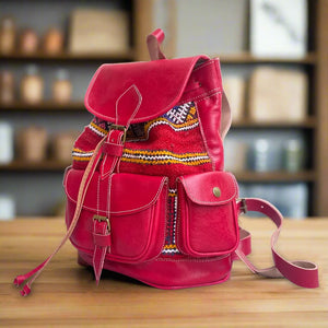 Angled side view of a red leather backpack adorned with colorful Moroccan kilim patterns, showcasing a spacious front pocket, two side pockets, and smooth leather straps, displayed on a wooden table