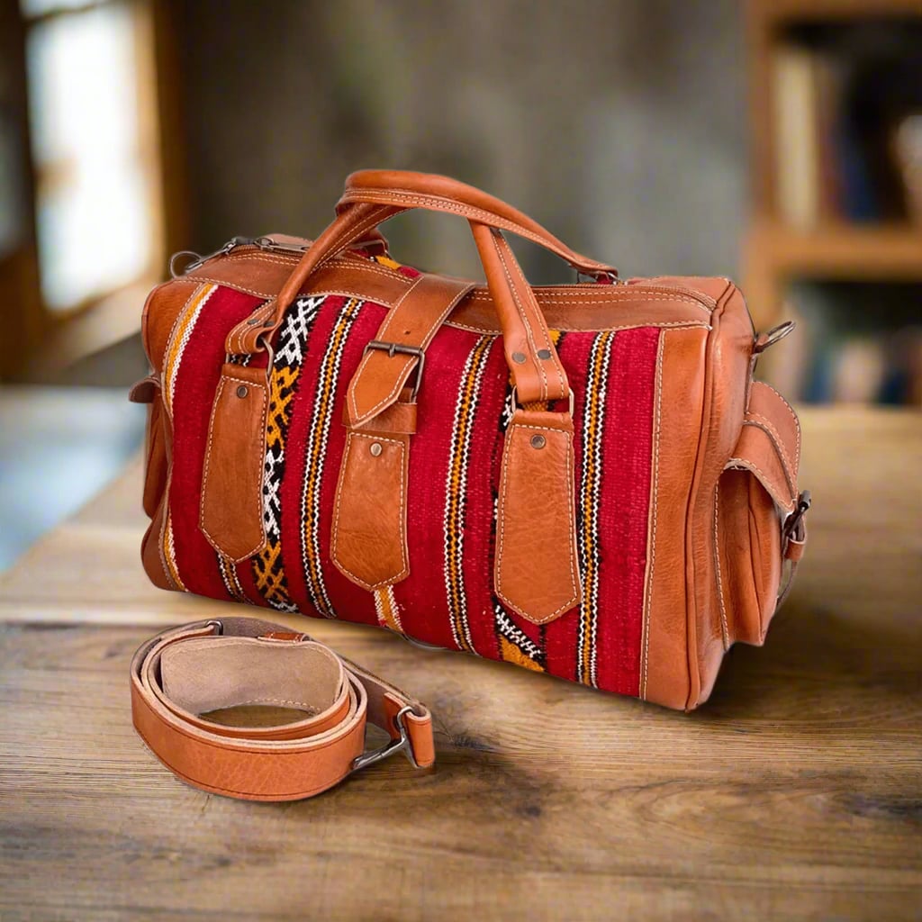 A handcrafted duffle bag featuring vibrant red handwoven kilim fabric with bold geometric patterns, accented by tan leather details. The bag includes sturdy stitched handles, a detachable leather shoulder strap, and metal hardware, resting on a wooden surface in a warm, rustic setting