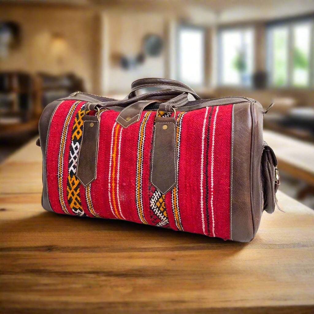 A leather and kilim duffle bag with intricate red, black, orange, and white geometric designs, complete with sturdy handles and spacious compartments, displayed on a wooden surface in a cozy interior