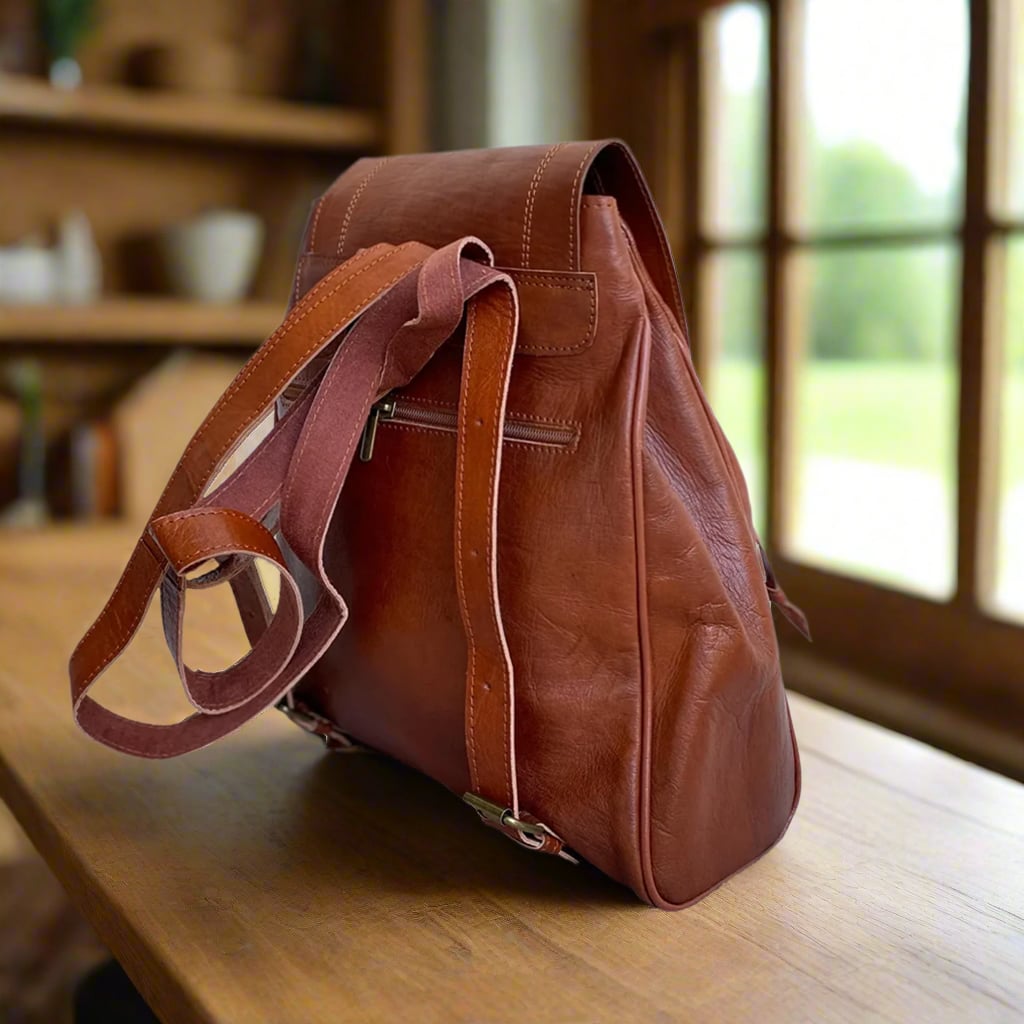 Back view of a brown leather backpack showcasing adjustable leather straps, a secure zippered back pocket, and a classic design, set near a window on a wooden surface