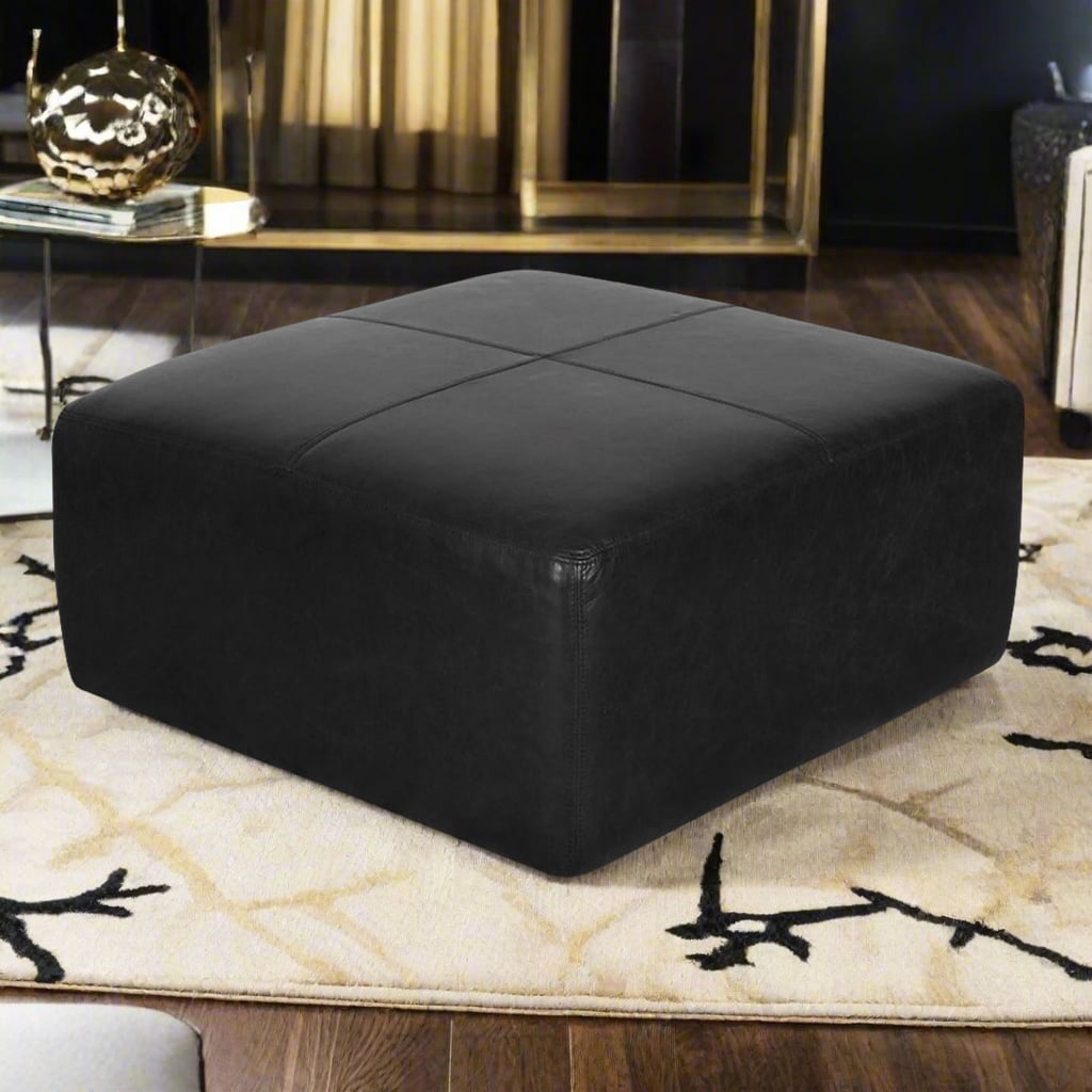 Handcrafted square black leather ottoman in a modern living room with a beige rug, golden decorative items, and a contemporary design.