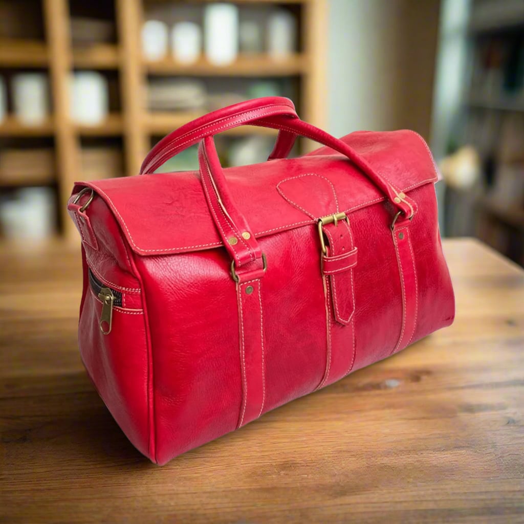 A bold red leather duffle bag crafted with premium leather, featuring a front buckle closure, dual reinforced stitched handles, and a spacious design. The bag includes a convenient side zip pocket with a brass zipper, precise stitching along the seams for durability, and an elegant yet practical structure. It is set on a smooth wooden surface in a warm interior, with soft lighting highlighting its vibrant color and fine craftsmanship