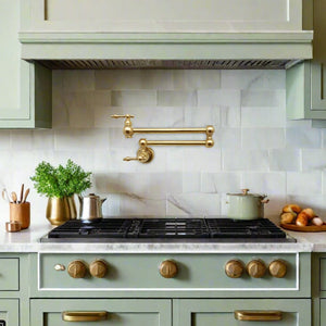 Brass wall-mount pot filler faucet installed above a stove in a traditional kitchen with green cabinetry, white marble backsplash, and gold hardware