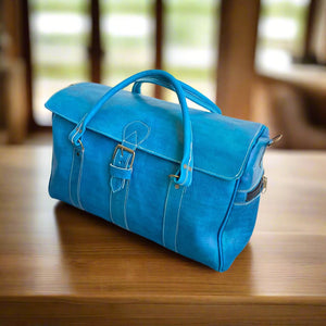 A vibrant blue leather weekend bag featuring a front buckle closure, double stitched handles, and a side zip pocket, resting on a polished wooden surface.