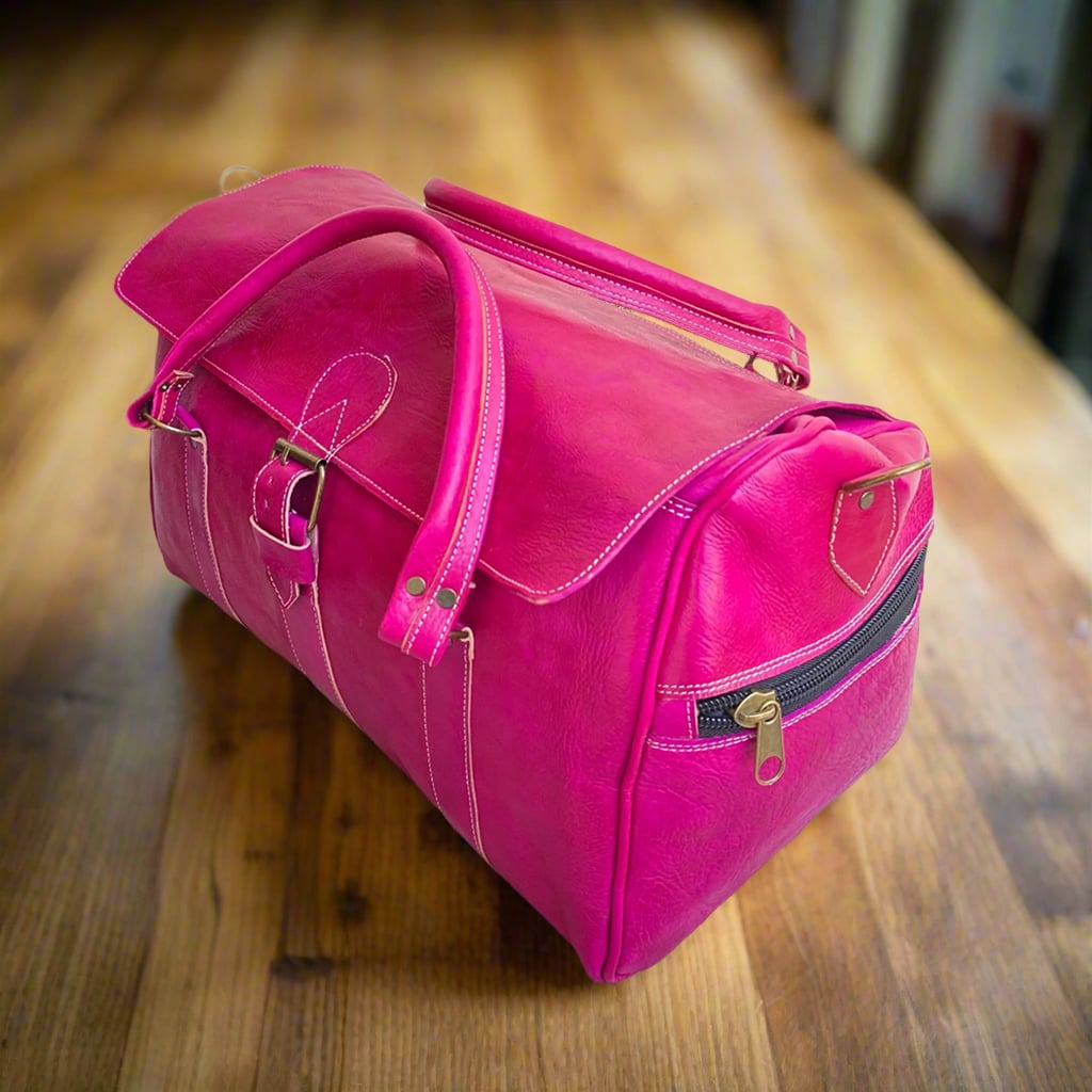 A vibrant pink leather duffle bag featuring a front buckle closure, sturdy stitched handles, and a side zip pocket, placed on a wooden surface in a warmly lit setting.
