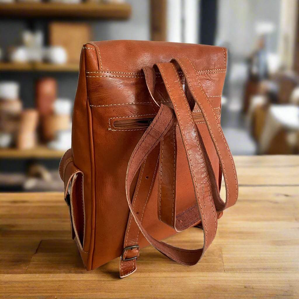 Back view of a structured brown leather backpack featuring adjustable leather straps, a secure zippered back pocket, and reinforced edges, set on a wooden surface in a rustic setting