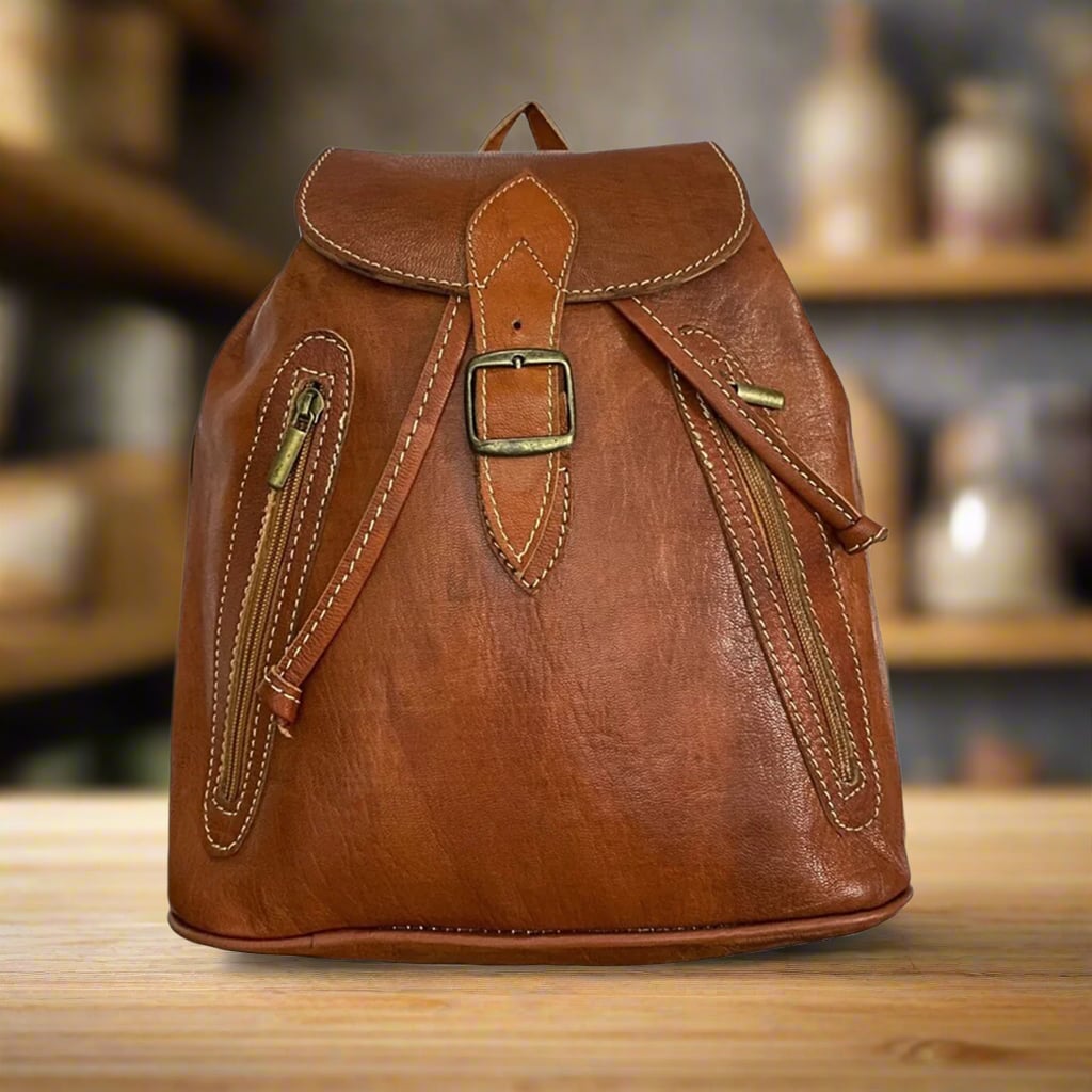 Front view of a brown leather backpack with a classic buckle closure, two vertical zippered side pockets, and detailed stitching, displayed on a wooden table in a cozy interior setting with pottery and shelves