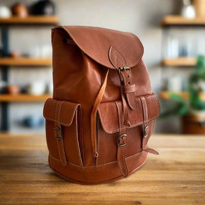 Side view of a handcrafted brown leather backpack showcasing two spacious buckle-closure side pockets, smooth adjustable leather straps, and precise hand-stitching, placed on a wooden table with shelves in the background