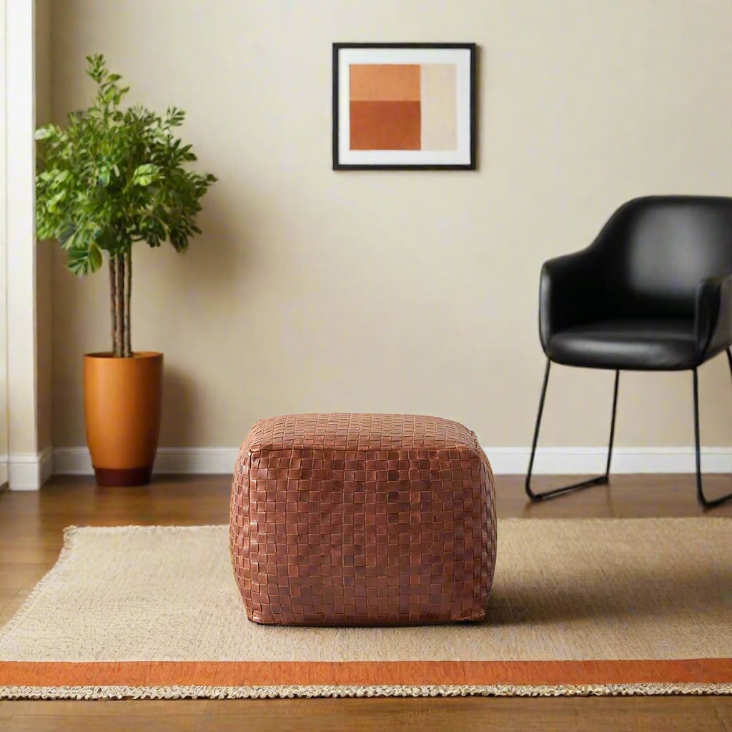 A square brown woven leather pouf sits on a beige and orange-trimmed rug in a modern living room. A black chair, potted plant, and abstract artwork decorate the space