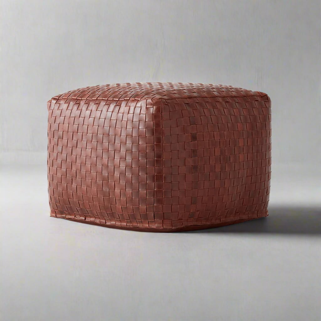 A square-shaped pouf made of brown woven leather sits on a plain gray surface with a neutral gray background. The leather has a tight basketweave pattern