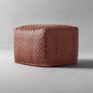 A square-shaped pouf made of brown woven leather sits on a plain gray surface with a neutral gray background. The leather has a tight basketweave pattern