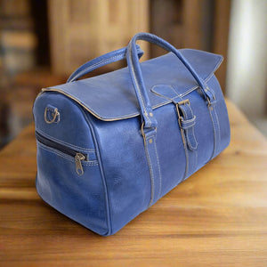 A classic blue leather duffle bag crafted with high-quality leather, featuring a front buckle closure, dual stitched handles, and a spacious design. The bag includes a convenient side zip pocket with a metallic zipper, reinforced seams for durability, and a sleek yet practical structure. It rests on a wooden surface in a warm, softly lit interior, highlighting its elegant craftsmanship and deep blue tone