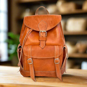 Front view of a classic tan leather backpack with buckle closures and side pockets, showcased on a wooden surface with bookshelves in the background