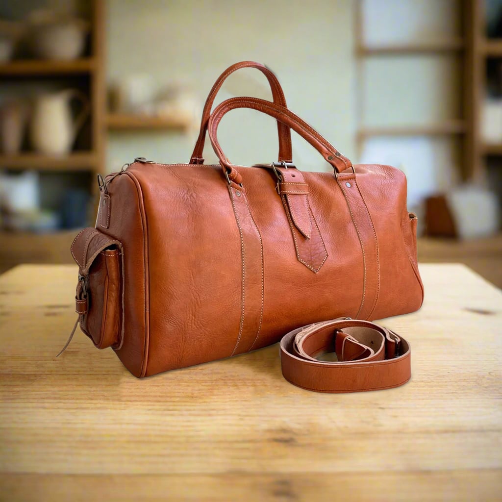 A classic tan leather duffle bag crafted from premium full-grain leather, featuring reinforced handles, a detachable shoulder strap, and a side pocket. The bag’s minimalist design highlights fine stitching and durable construction, resting on a wooden surface in a warm, softly lit setting