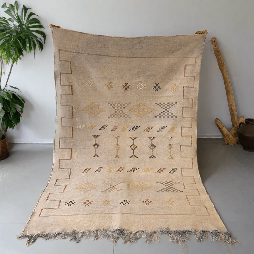 Handwoven cream Moroccan rug featuring traditional Berber motifs in earthy tones, with geometric borders and fringe detail, displayed indoors