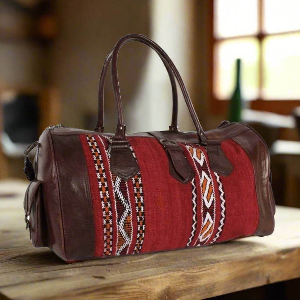 A handcrafted duffle bag made with dark brown leather accents and sturdy handles, paired with vibrant red kilim fabric showcasing intricate geometric patterns in black, white, and orange. The bag features side pockets, reinforced stitching, and a classic rectangular shape, blending traditional craftsmanship with modern functionality. Set on a rustic wooden table with a softly lit background.