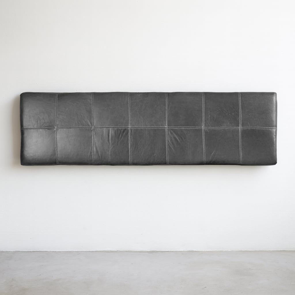 A full view of a wall-mounted black leather headboard with grid-style stitching, set against a clean white wall and concrete floor