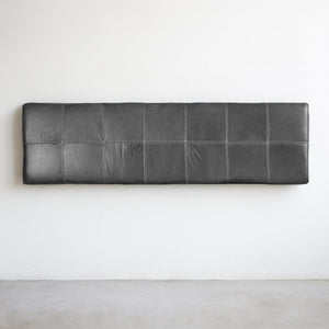 A full view of a wall-mounted black leather headboard with grid-style stitching, set against a clean white wall and concrete floor