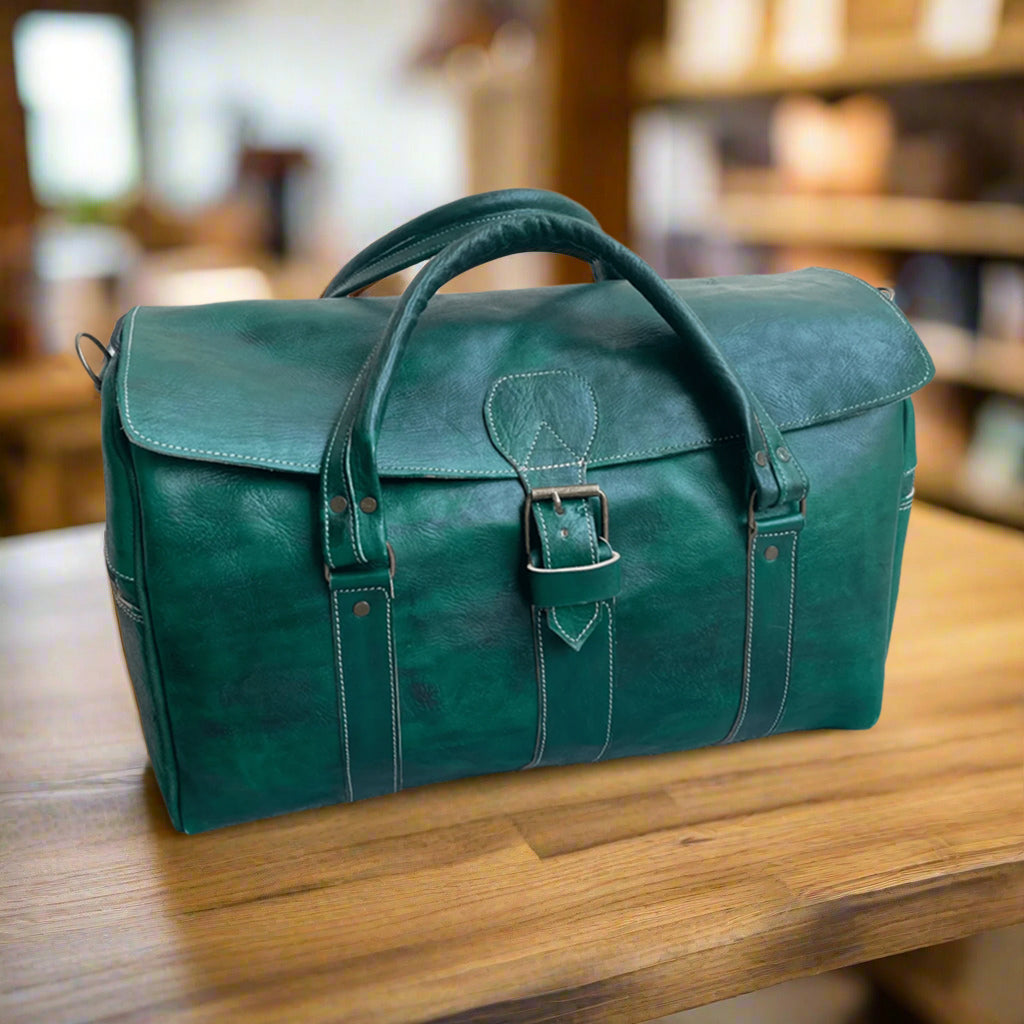 A deep green leather duffle bag featuring a front buckle closure, reinforced stitched handles, and a sleek design, placed on a polished wooden surface in a softly lit setting.