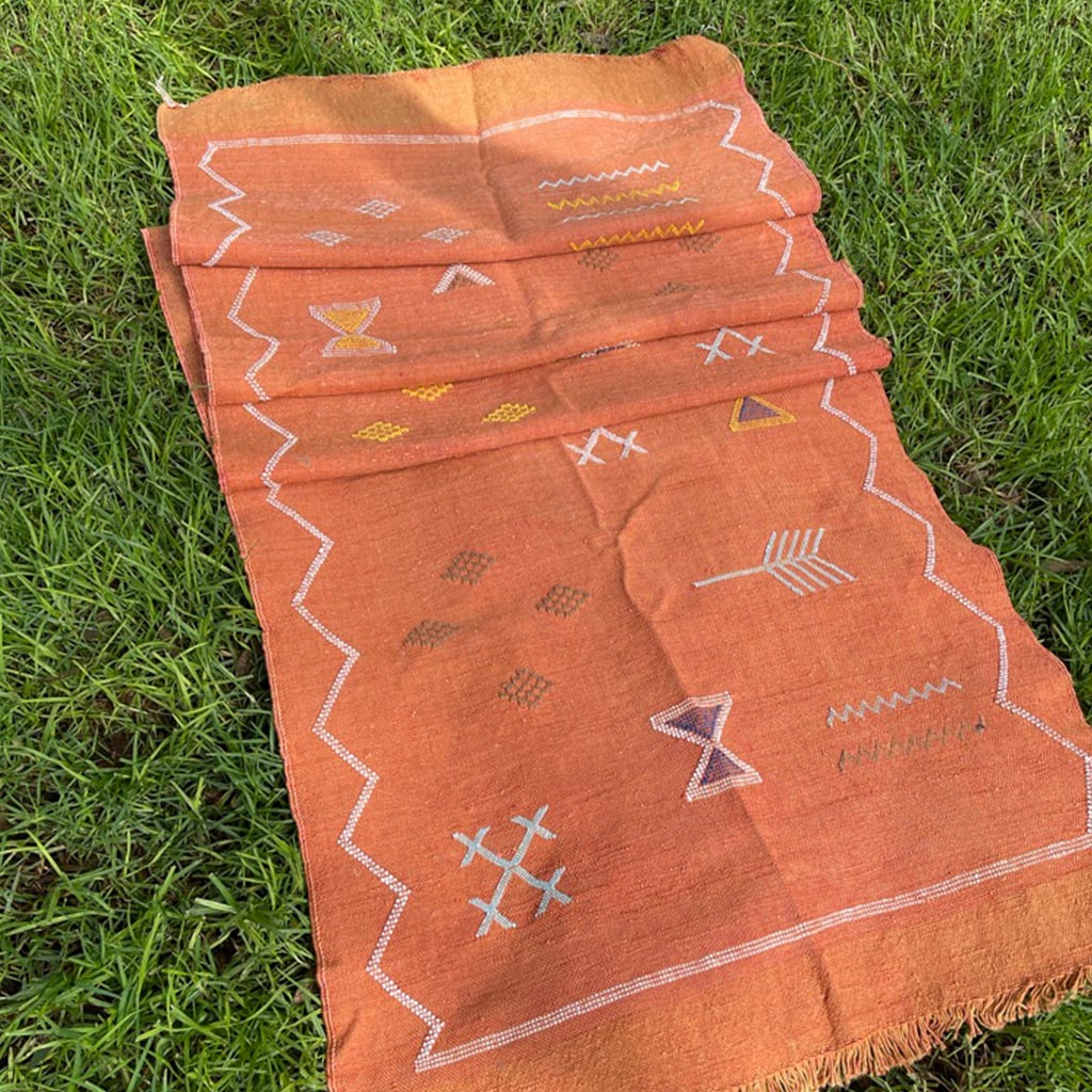 Folded view of a terracotta Moroccan runner rug with geometric embroidery and white zigzag borders, placed on green grass