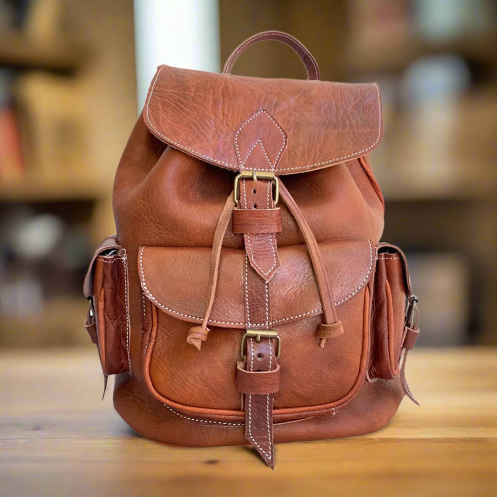 Front view of a handmade brown leather backpack with a buckle-closure flap, a spacious front pocket, and two side pockets, placed on a wooden table in a cozy setting