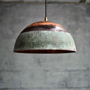 Artisanal green patina and copper pendant light hanging near a window, casting warm ambient light in a textured setting