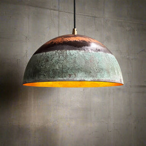 handcrafted dome pendant light with green patina and copper top finish, glowing warmly against a concrete background