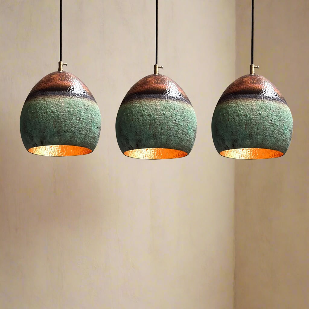 Three hammered copper pendant lights with green patina finish hanging in a row against a warm beige wall.