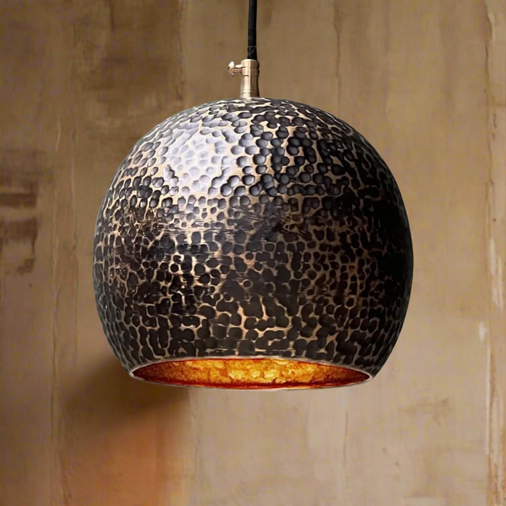 Close-up of a hammered black brass globe pendant light with a warm inner glow, hanging in front of a textured beige wall