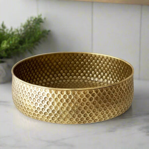 Side view of a hammered brass round basin with dotted texture, sitting on a marble surface with a plant in the background