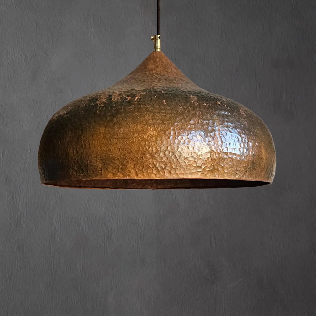 Hand-hammered copper pendant lamp with a wide dome-shaped shade, suspended from a black cord and brass fitting, against a dark gray background