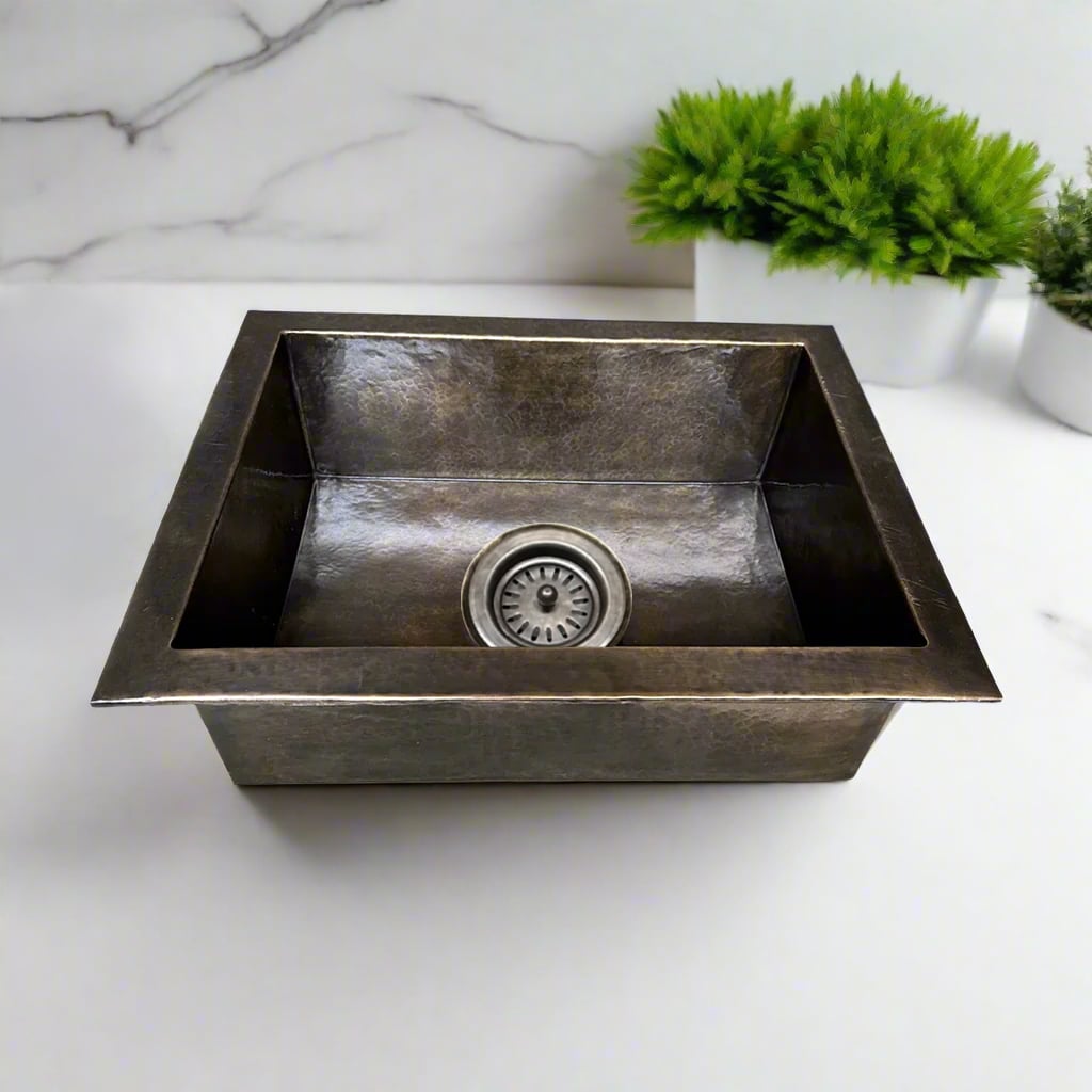 Square hammered nickel undermount kitchen sink with a hammered texture, placed on a white countertop with green plants in the background