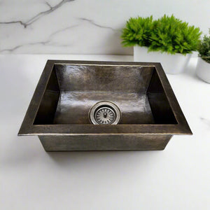 Square hammered nickel undermount kitchen sink with a hammered texture, placed on a white countertop with green plants in the background