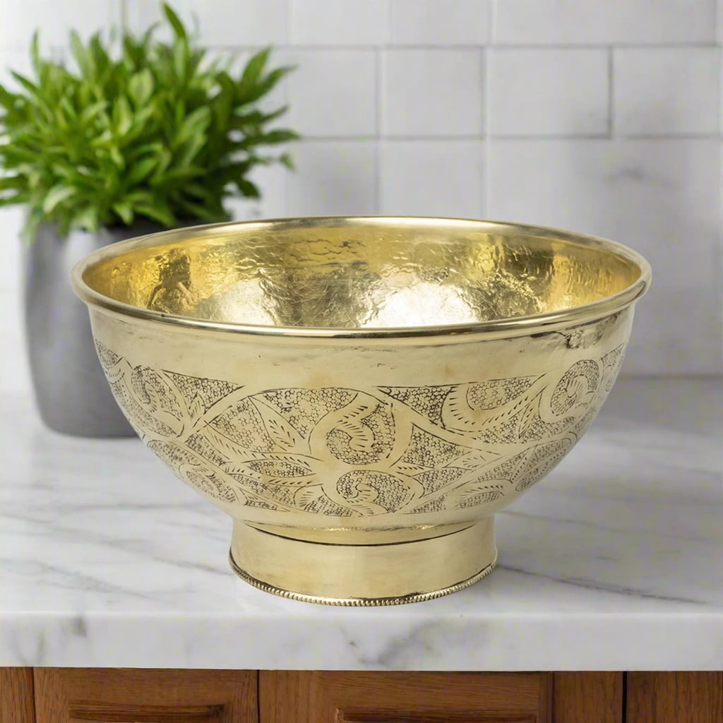 Round brass bathroom sink with hammered interior and ornate embossed exterior, placed on a marble countertop