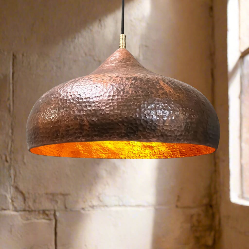 Hand-hammered copper pendant light with a warm glowing interior, hanging in front of a textured plaster wall and soft natural light