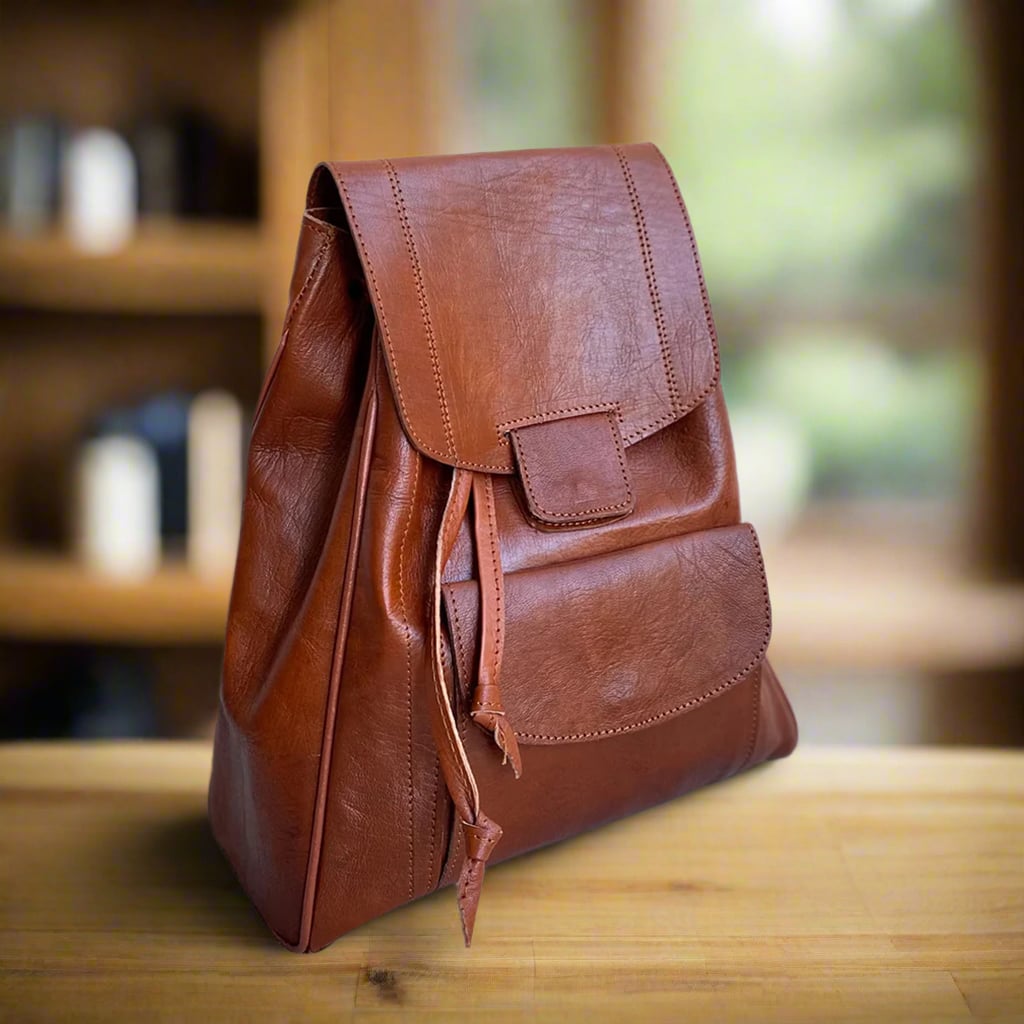 Angled front view of a handcrafted brown leather backpack featuring a flap closure with a front pocket, sleek stitching, and a smooth finish, displayed on a wooden table