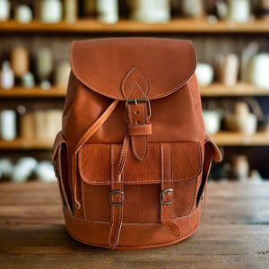 Front view of a brown leather backpack with a classic buckle-closure flap, two roomy front pockets with buckle straps, and fine hand-stitched detailing, displayed on a wooden table against a backdrop of pottery and shelves