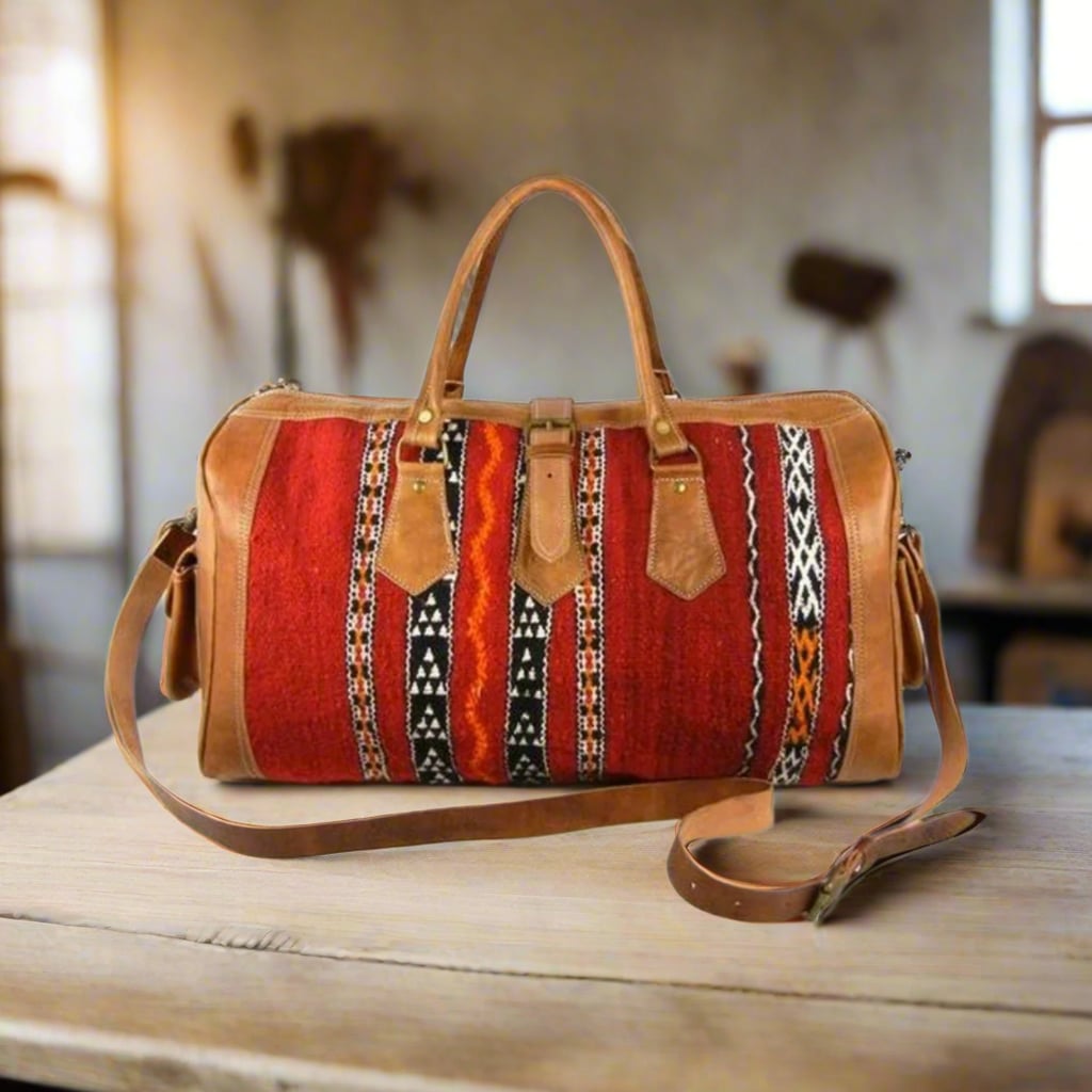 A handcrafted duffle bag featuring premium tan leather panels and accents, combined with vibrant red handwoven kilim fabric adorned with intricate geometric patterns. The bag includes sturdy double leather handles with reinforced stitching, a detachable and adjustable leather shoulder strap, and a structured rectangular shape. It rests on a wooden surface in a rustic setting with warm, natural lighting.