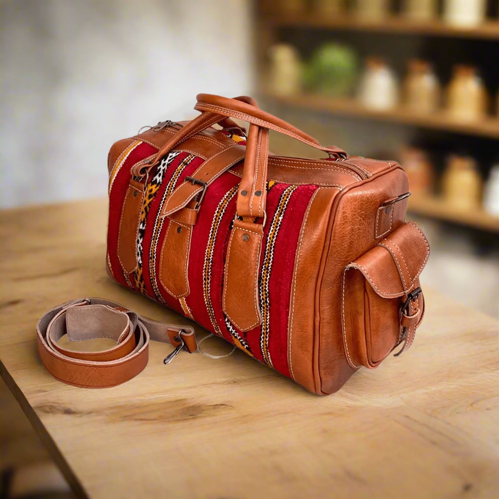 An artisan duffle bag crafted from vibrant red kilim fabric with intricate geometric designs, complemented by tan leather accents. The bag features side leather pockets, reinforced handles, and a detachable strap, displayed on a wooden table in a cozy, softly lit interior