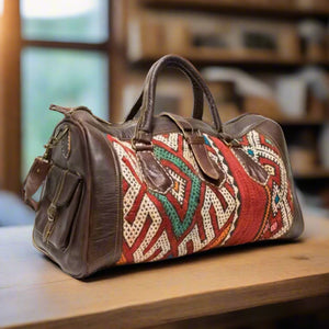 A three-quarter view of a Moroccan travel bag featuring premium brown leather and bold kilim details in red, green, and white geometric patterns