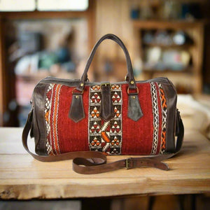 Handcrafted leather and kilim duffle bag featuring vibrant red and multicolored handwoven kilim panels with dark brown leather trim, adjustable strap, and sturdy handles, placed on a wooden surface in a rustic interior setting