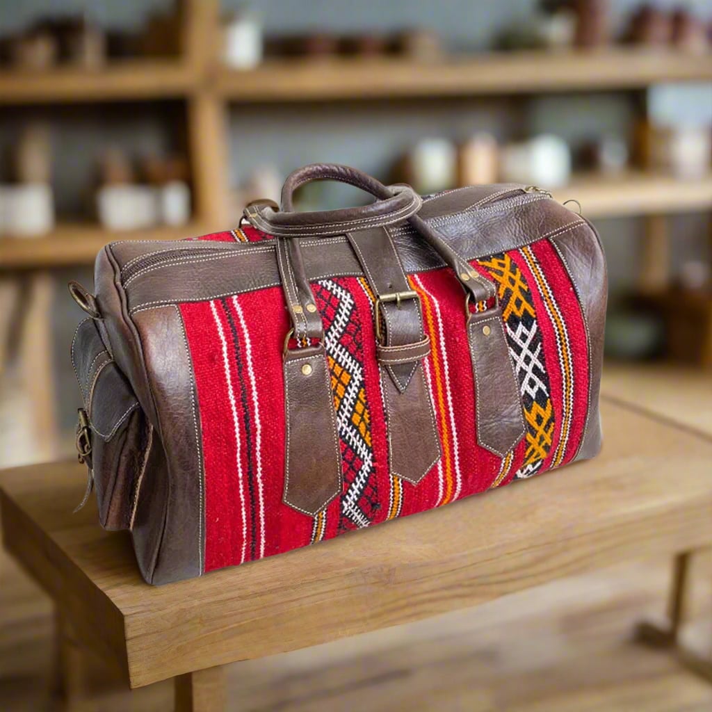 A handcrafted duffle bag featuring rich brown leather accents and vibrant red kilim fabric with bold geometric patterns, resting on a wooden table in a warm setting.