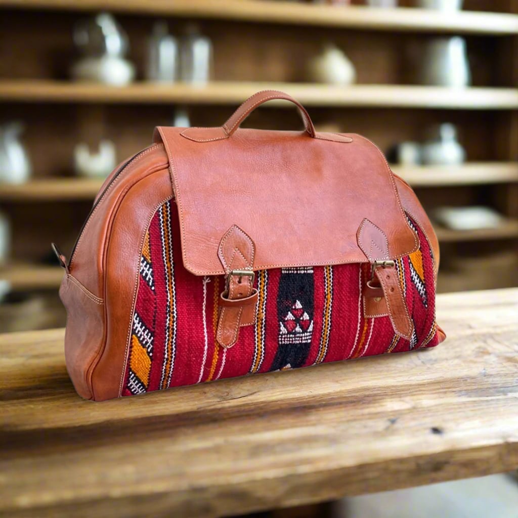 A handcrafted travel bag featuring tan leather accents and vibrant red kilim fabric with intricate geometric patterns, secured with dual buckle closures. Displayed on a wooden table in a cozy, softly lit setting