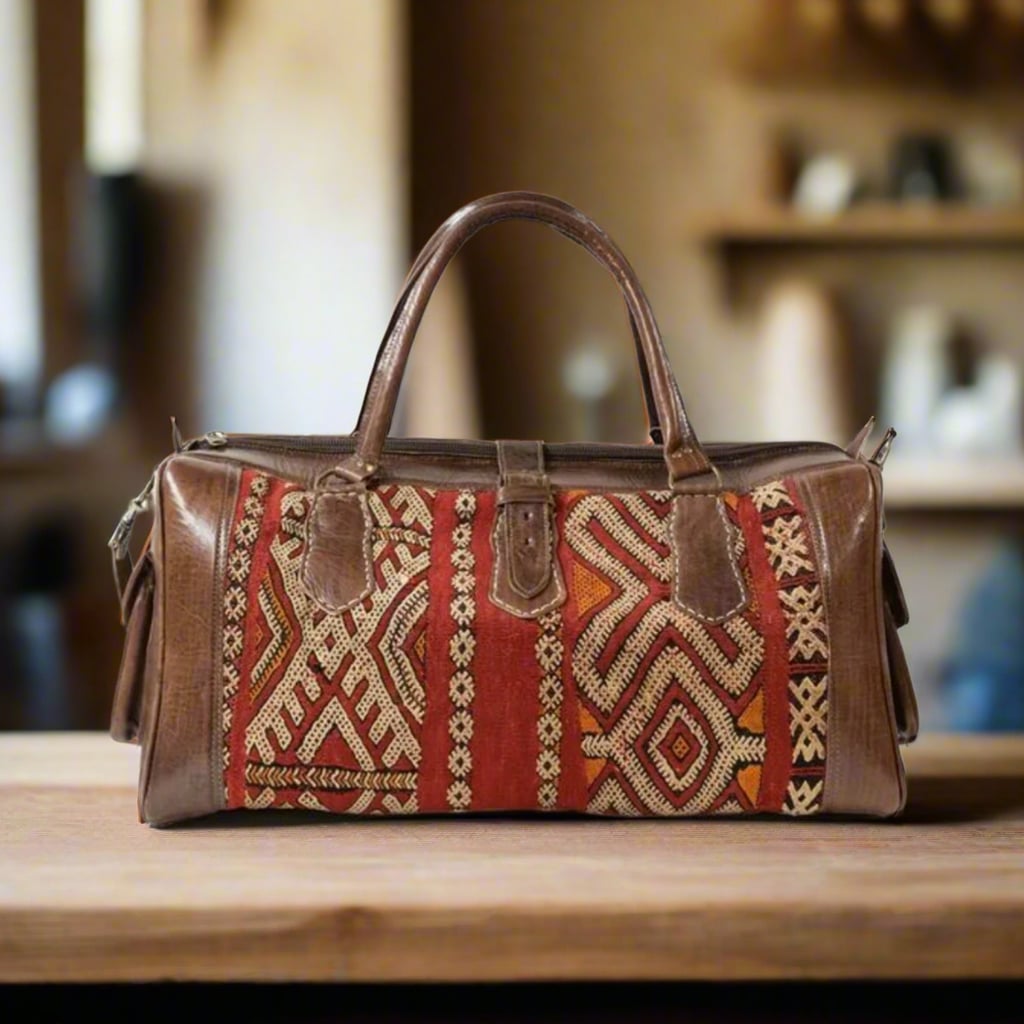 A handcrafted leather and kilim travel duffle bag featuring intricate traditional Moroccan patterns in vibrant red, orange, and beige tones, with sturdy leather straps and side pockets, set on a wooden surface in a cozy interior