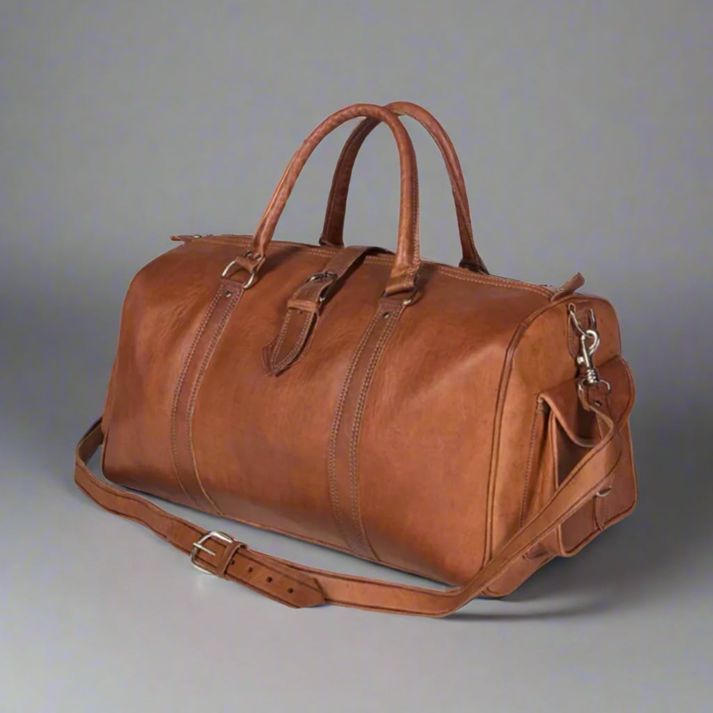 A side view of a handcrafted leather travel bag displayed against a plain background. The bag is made of warm tan leather, featuring double-stitched detailing, reinforced carry handles, an adjustable shoulder strap with a silver buckle, and a side pocket secured with a leather strap. Its structured yet soft design makes it perfect for travel, whether for a weekend getaway or a business trip