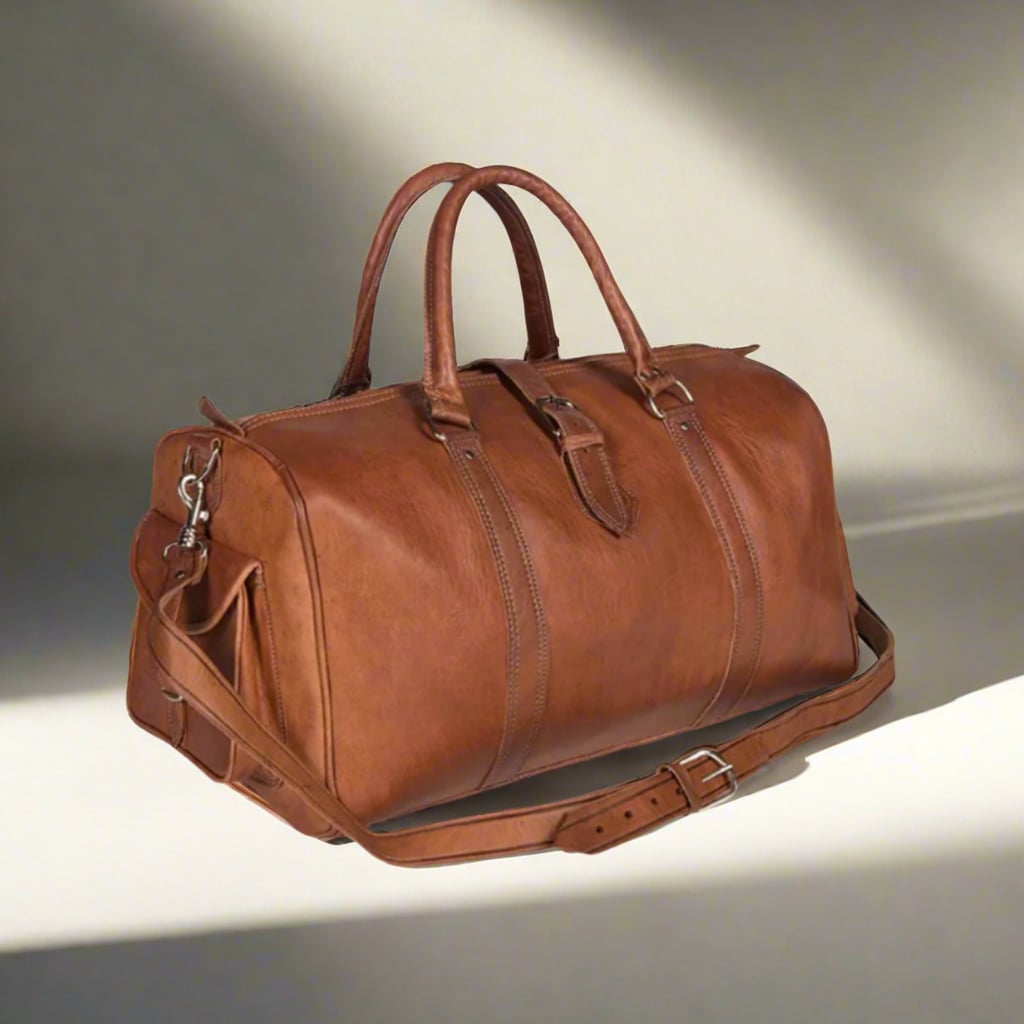 A three-quarter view of a premium leather travel bag resting in soft, natural light, emphasizing its rich tan color and smooth, supple texture. The bag features sturdy handles, a detachable shoulder strap with silver hardware, and a streamlined silhouette with a small side pocket and buckle for added functionality. Ideal for travelers seeking durability and elegance in one stylish accessory