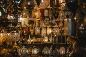 A display of handcrafted Moroccan brass lanterns and lamps with intricate pierced designs, casting warm light and decorative patterns in various shapes and sizes